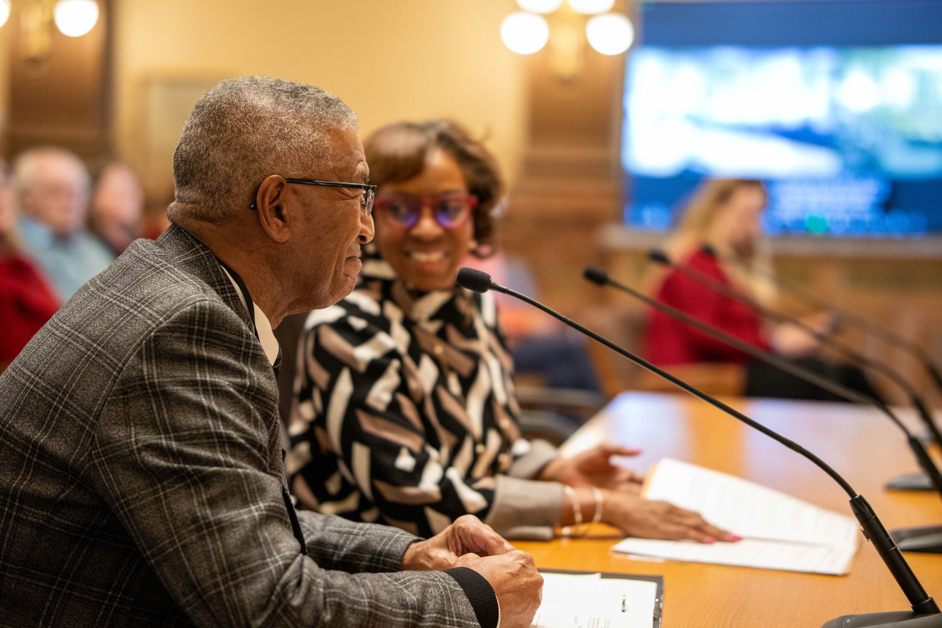 Two people speaking at a conference table with microphones in a meeting room