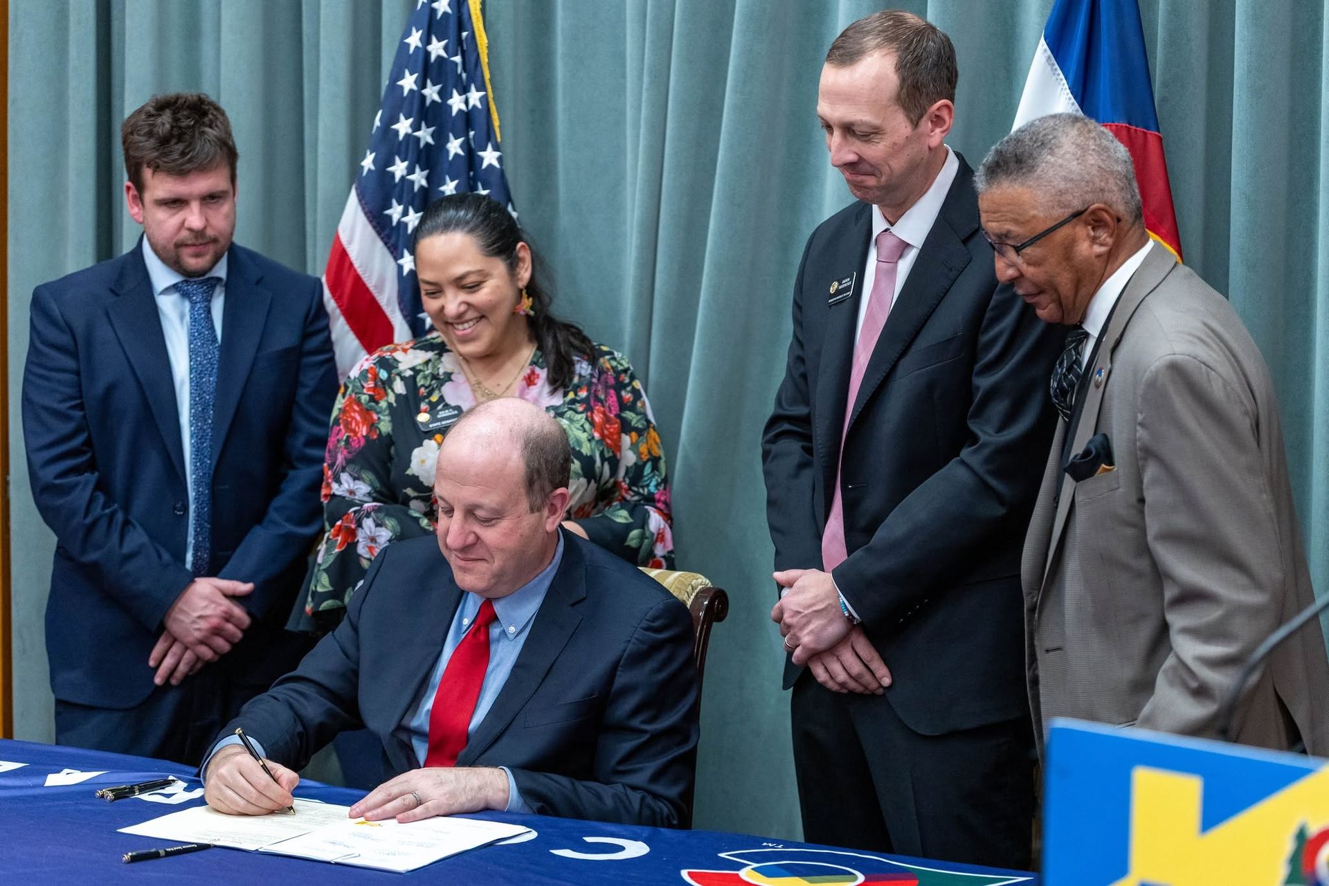 Man signing a document at a table while four others stand behind him in front of U.S. and Texas flags