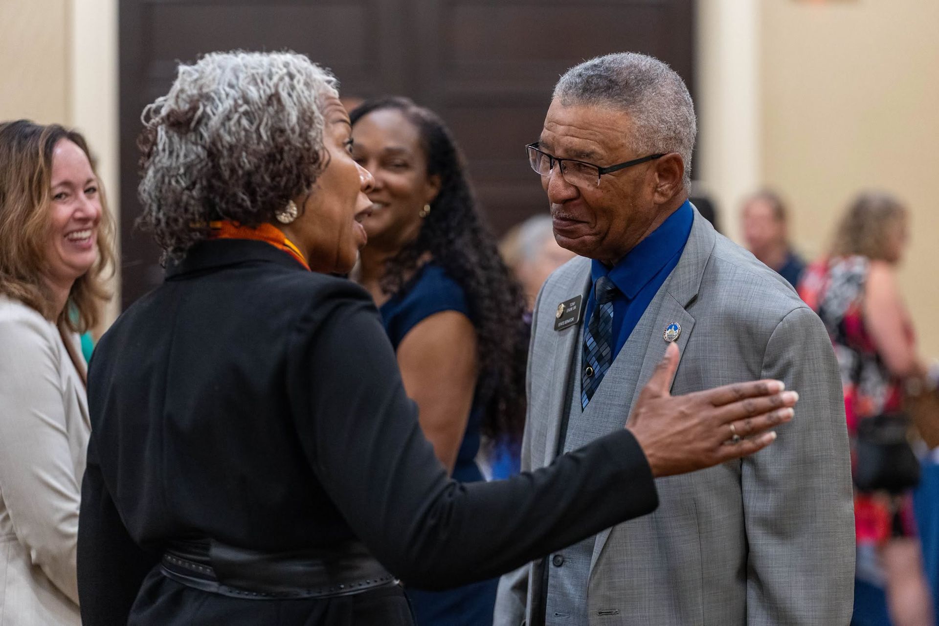 Two people smiling and greeting each other at an indoor social event.