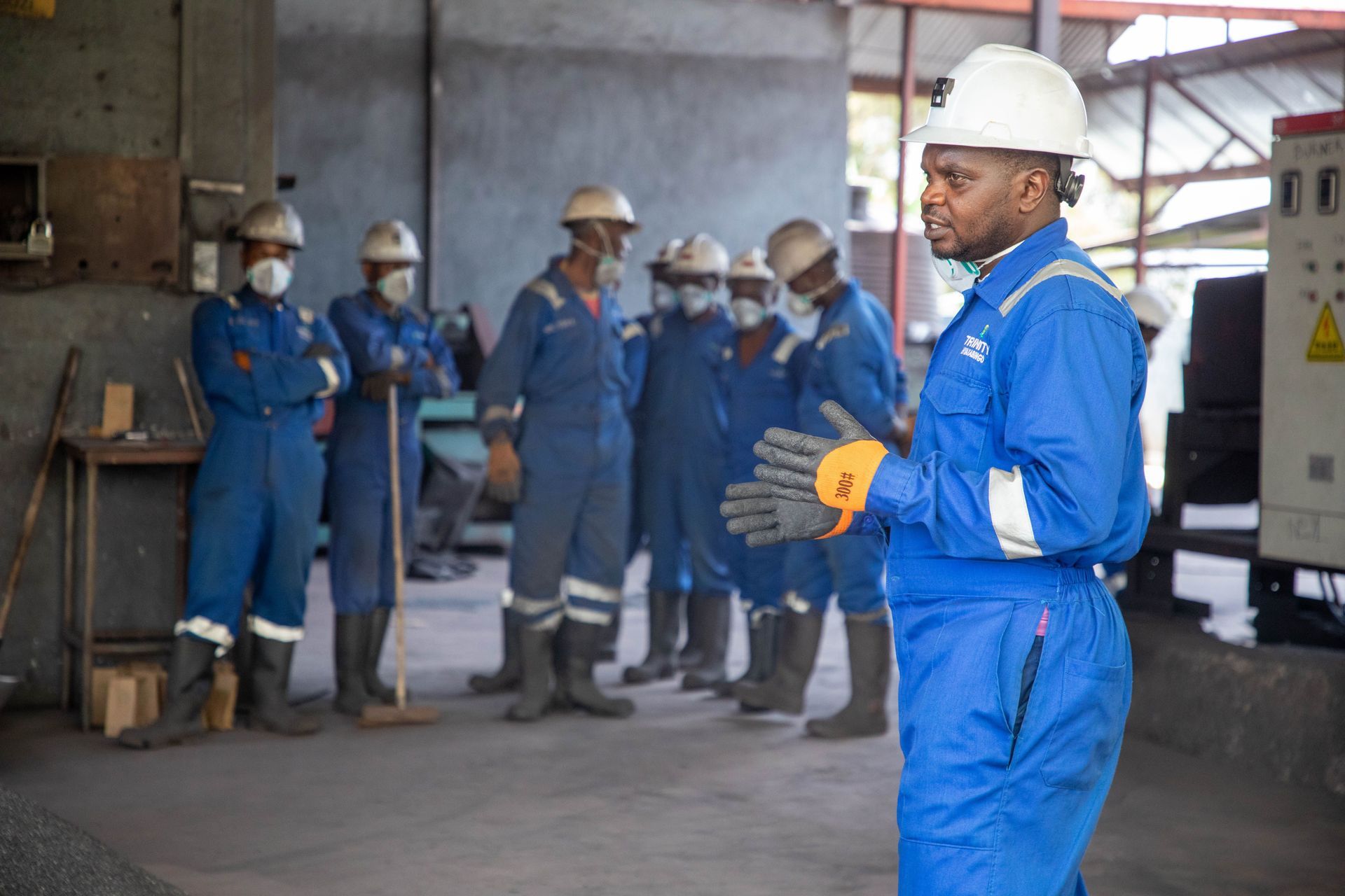 Workers in blue coveralls and hard hats standing in an industrial warehouse, one in the foreground speaking.