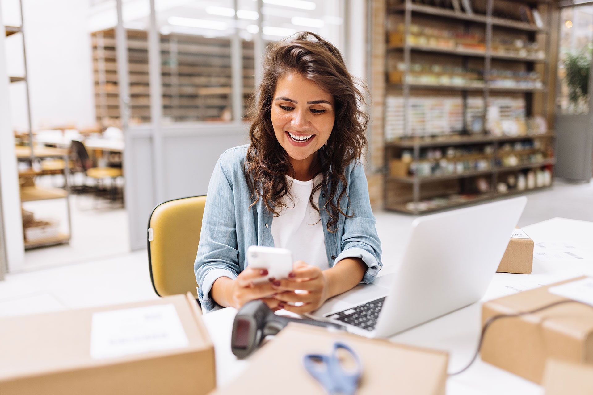 A woman is sitting at a desk with a laptop and a cell phone.