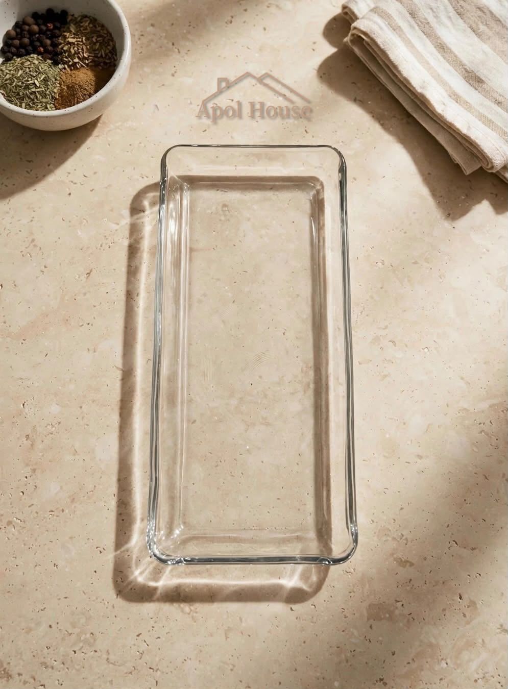 Una fuente rectangular de vidrio vacía sobre una encimera beige, con cuencos y un paño doblado cerca