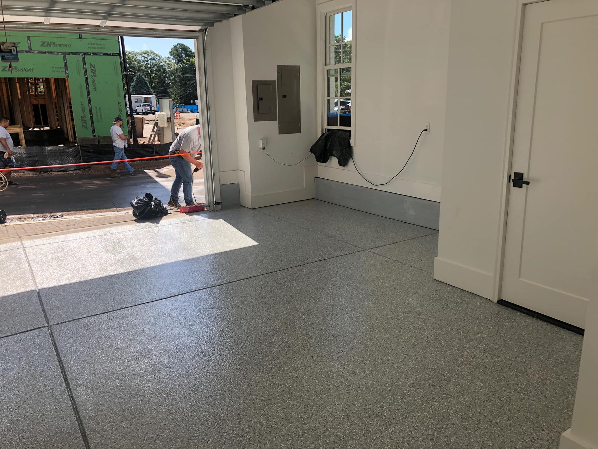 Garage interior with gray speckled epoxy floor, white walls, and a door. Construction workers visible outside.