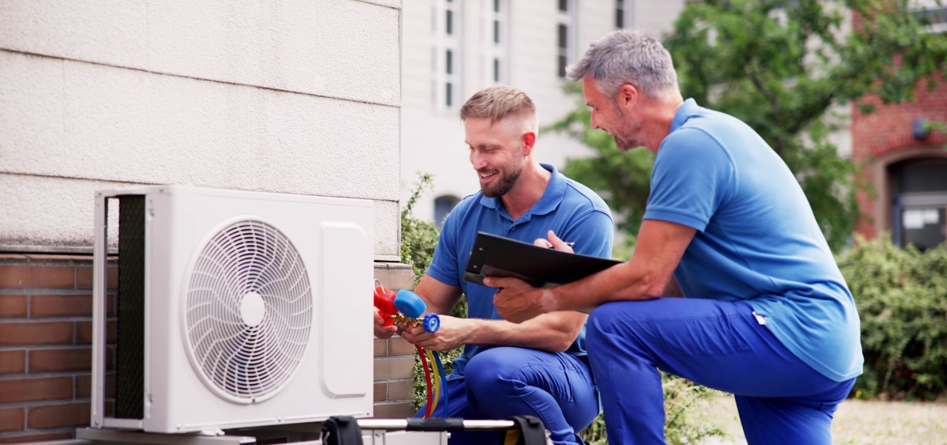 Two technicians in blue uniforms work together to inspect an outdoor HVAC unit, with one checking a clipboard.