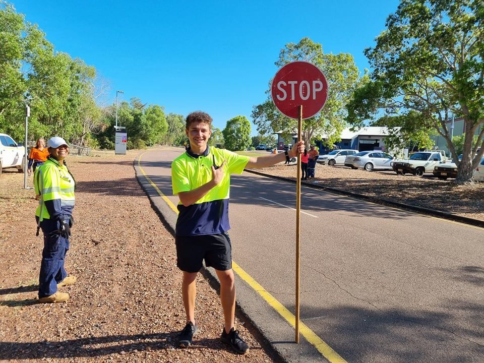 Two road workers: one holds a stop sign, the other stands near a road, both are wearing high-visibility vests.
