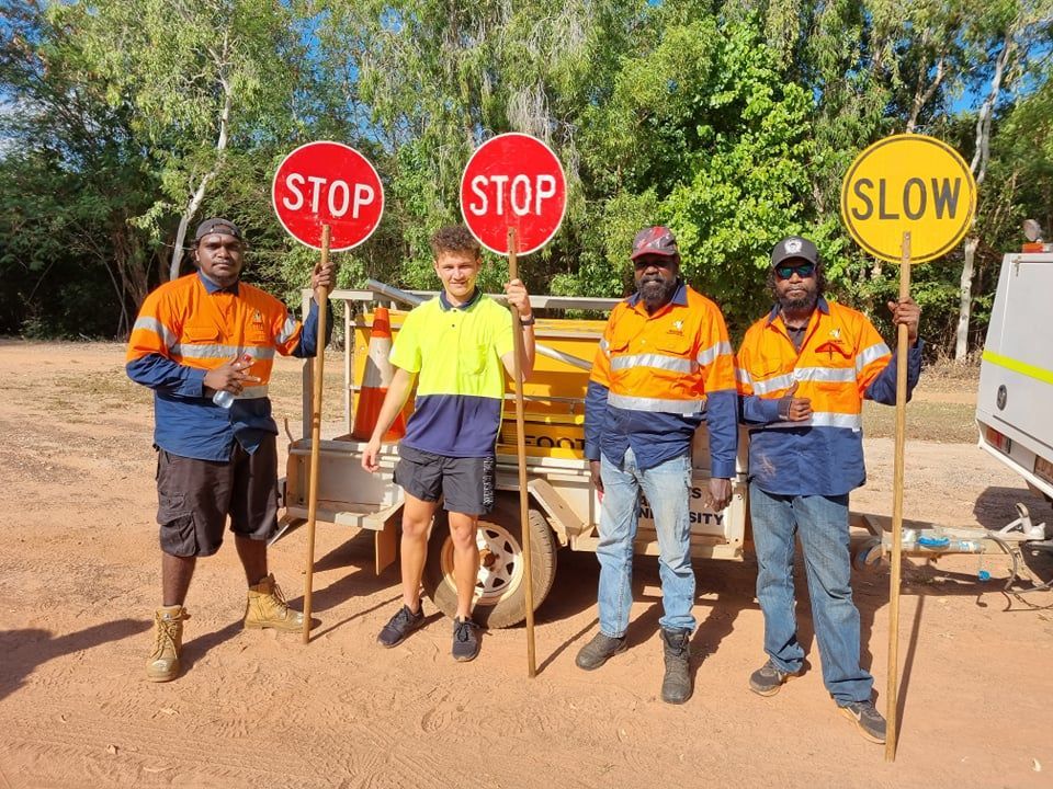 Four road workers holding stop and slow signs near a trailer in a sunny outdoor setting.