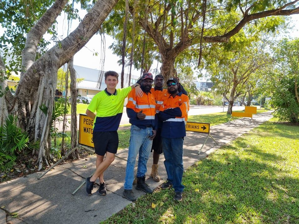 Four construction workers standing together on a sidewalk, posing under a tree; one in yellow, three in orange.