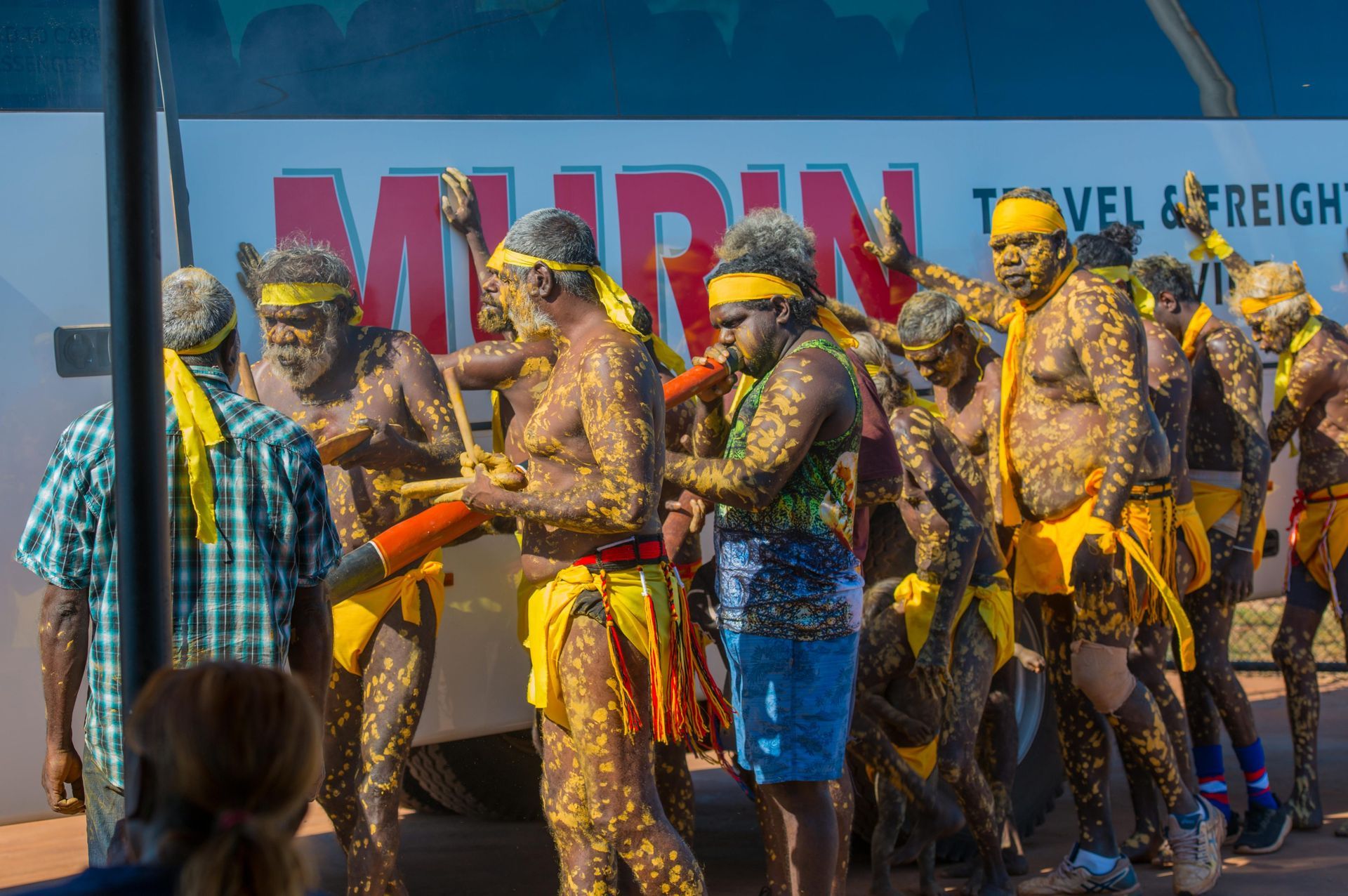 Indigenous Australians performing a traditional ceremony outdoors. Men wear body paint, yellow cloth, and play didgeridoos.