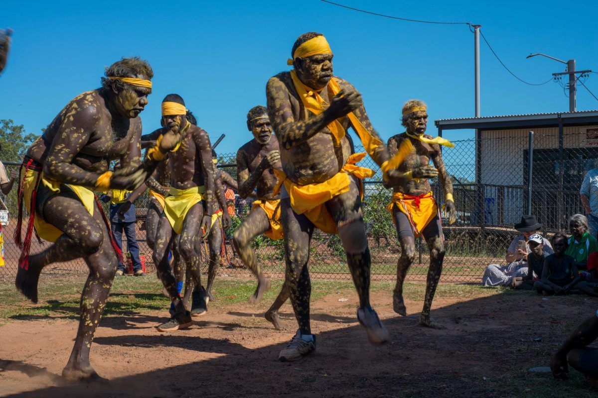Aboriginal Australians in yellow and brown body paint perform a traditional dance outdoors.