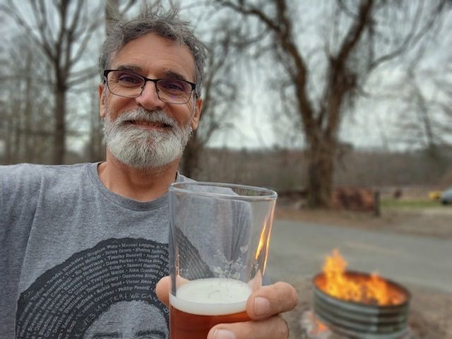 Man holding a beer glass outdoors beside a fire pit with flames in the background