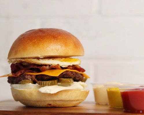 Burger with egg, bacon, and cheese on a brioche bun, with three dipping sauces, on a wooden board.