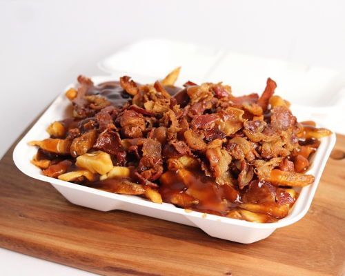 Poutine with bacon in a white container on a wooden board. Fries topped with gravy and crispy bacon.