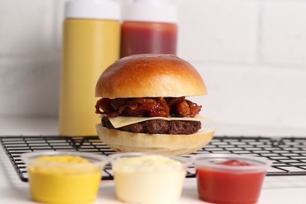 Burger with cheese and bacon on a bun, with mustard and ketchup bottles and small condiment cups.