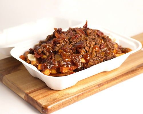 Fries in a white takeout container, topped with meat and sauce, on a wooden board.