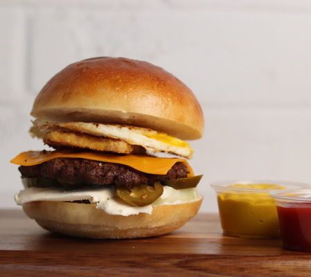 Burger with egg, cheese, jalapeños, and sauce on a wooden surface, with mustard and ketchup.