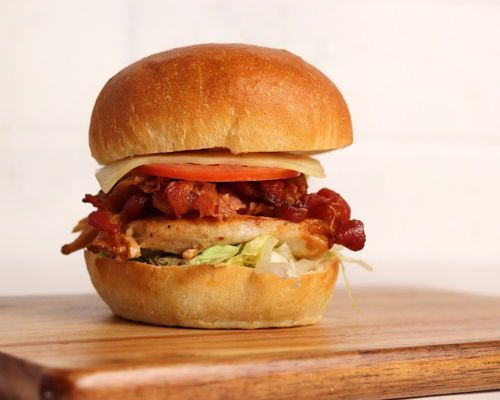 Chicken sandwich with bacon, tomato, cheese, and lettuce on a toasted bun, resting on a wooden board.