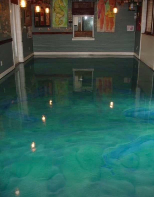 A room with a shiny, turquoise-colored epoxy floor. The walls are a dark grey, with abstract art. A room with a shiny, turquoise-colored epoxy floor. The walls are a dark grey, with abstract art.
