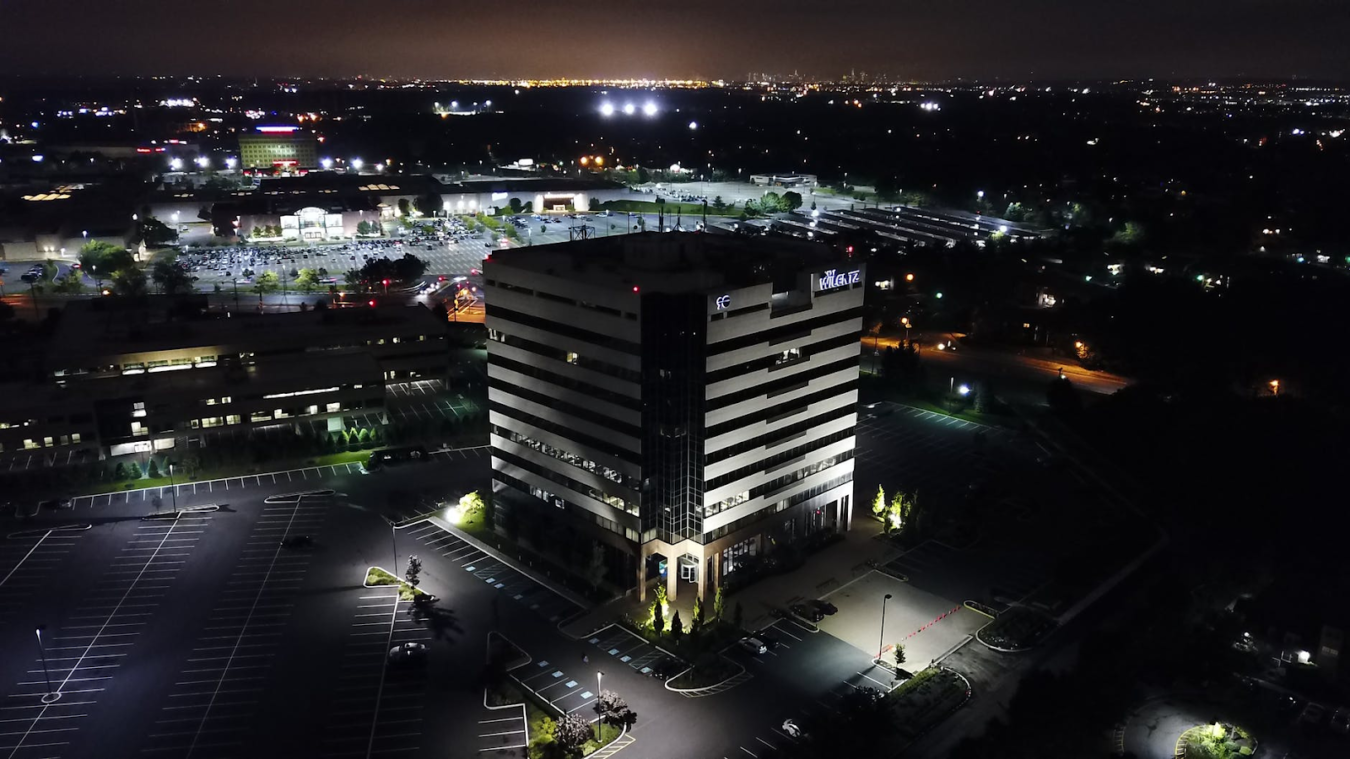 Night aerial view of a lit office building with parking lots and city lights in the background