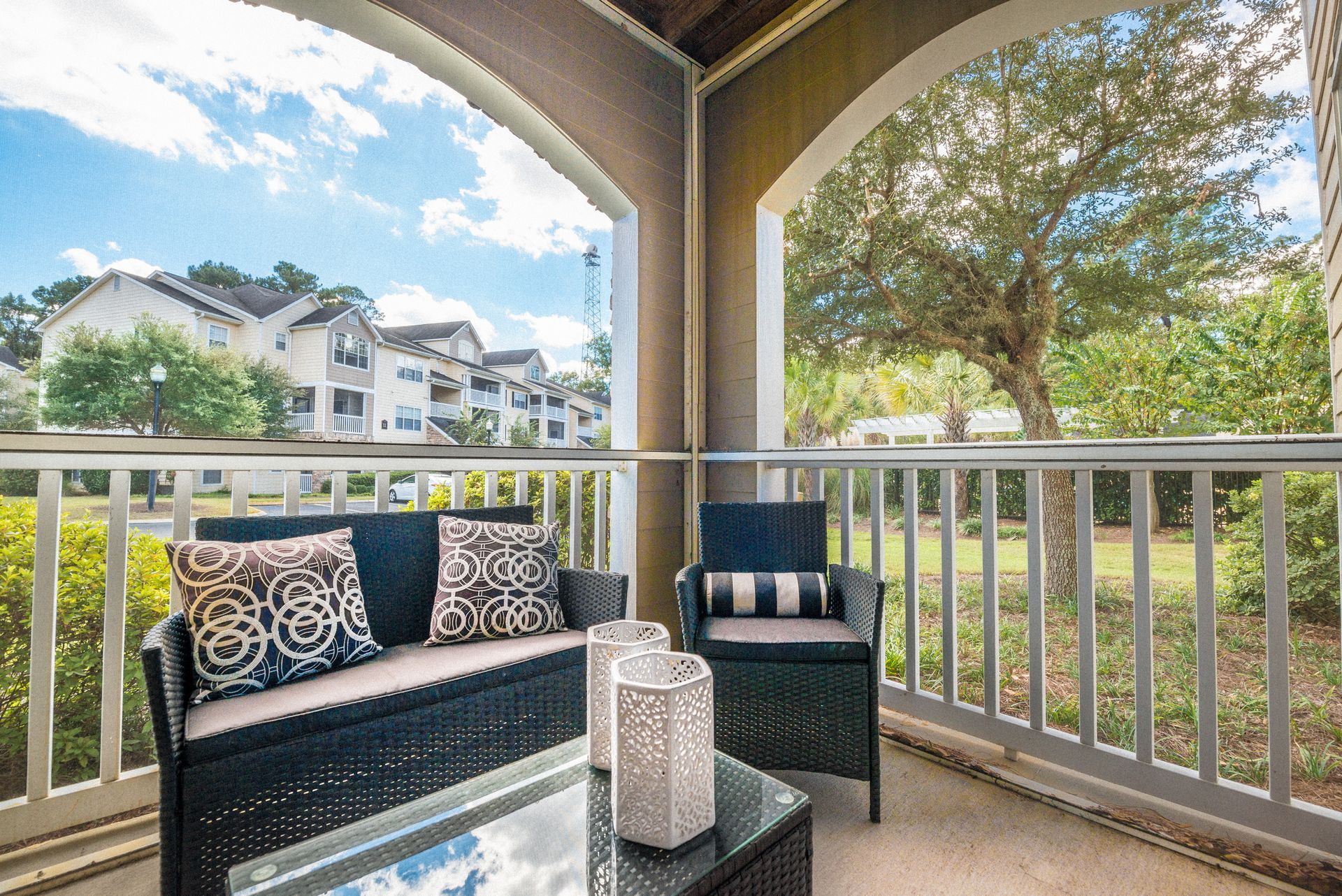 Covered balcony with black wicker seating, lantern table, and a view of townhomes and trees at Walden at Chatham Center Apartments in Savannah, GA.