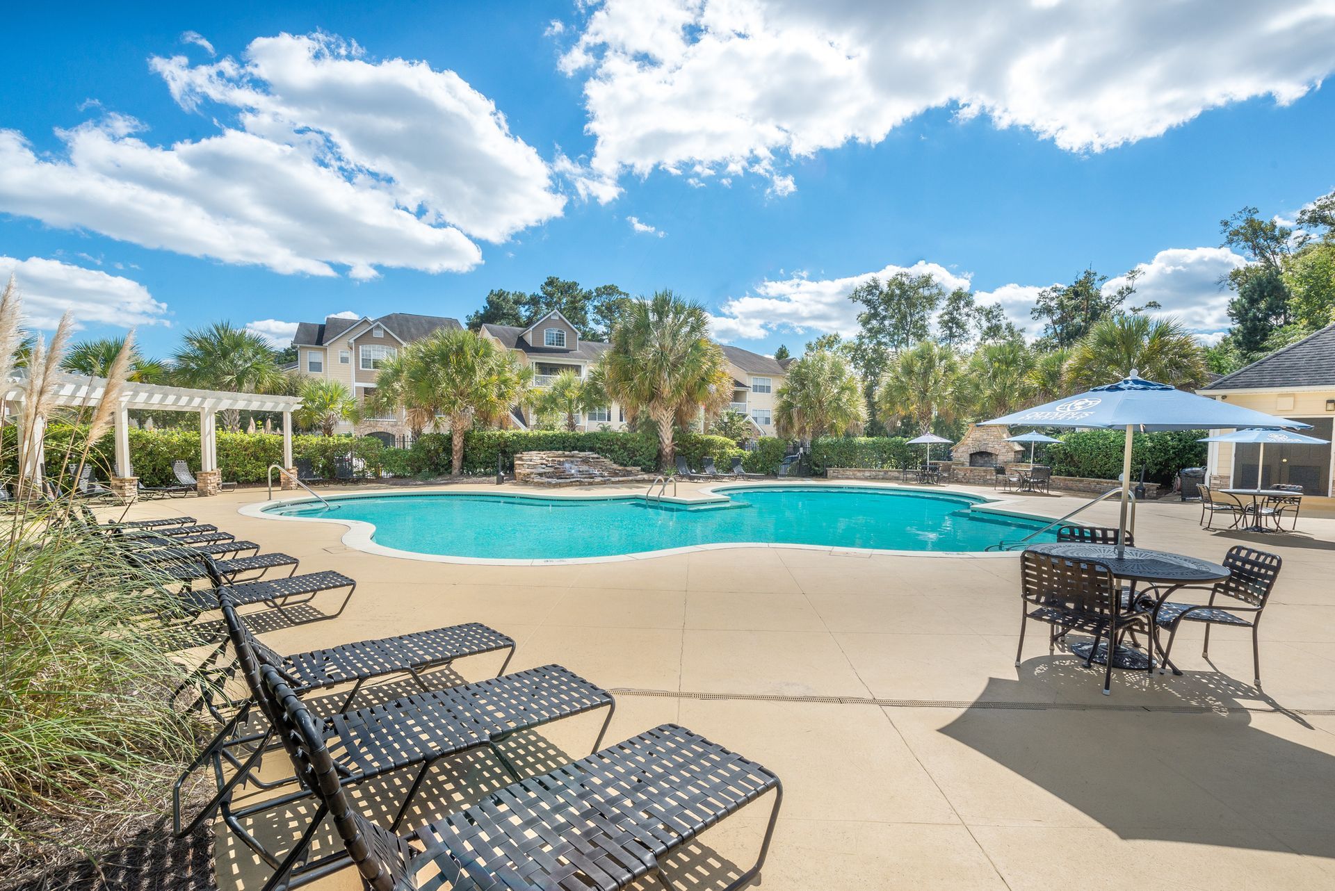 Resort pool with lounge chairs, umbrellas, and palm trees under a bright blue sky at Walden at Chatham Center Apartments in Savannah, GA.