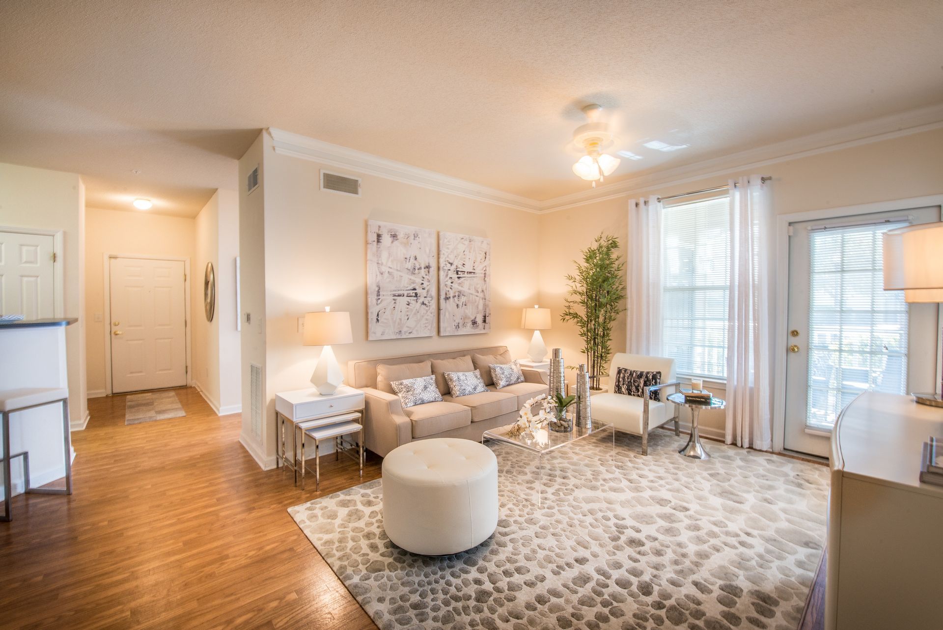 Bright living room with beige sofa, patterned rug, ottoman, plants, and sunlight through windows and blinds at Walden at Chatham Center Apartments in Savannah, GA