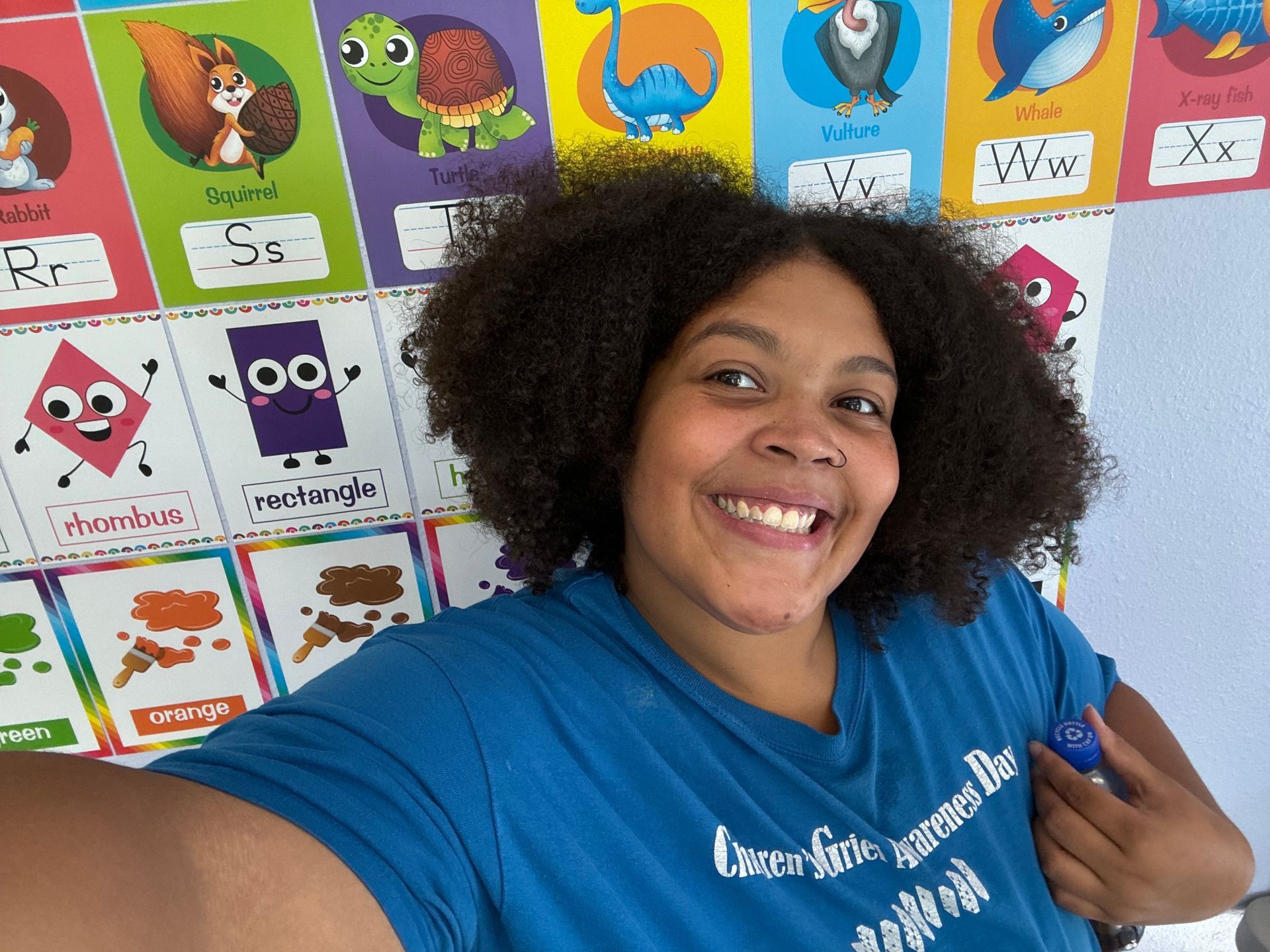 A woman in a blue shirt is taking a selfie in front of an alphabet poster.