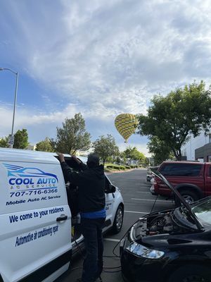 A man is working on a car in a parking lot with a hot air balloon in the background.