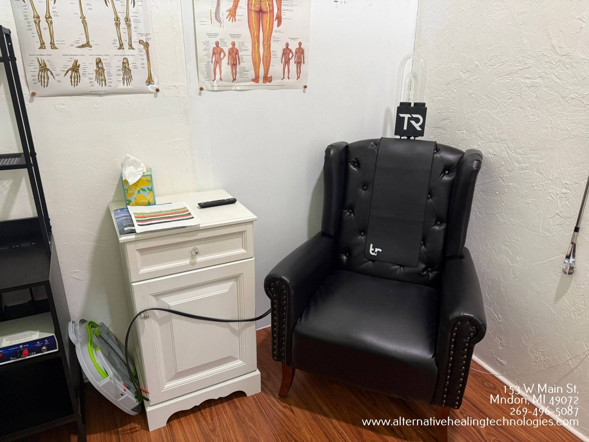 A black armchair with a headrest in a medical office. White cabinet with items, anatomical charts on the wall.