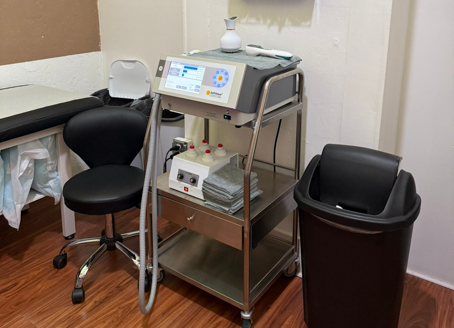 Medical office exam room with black stools, rolling cart, exam table, and wall-mounted tissue holder