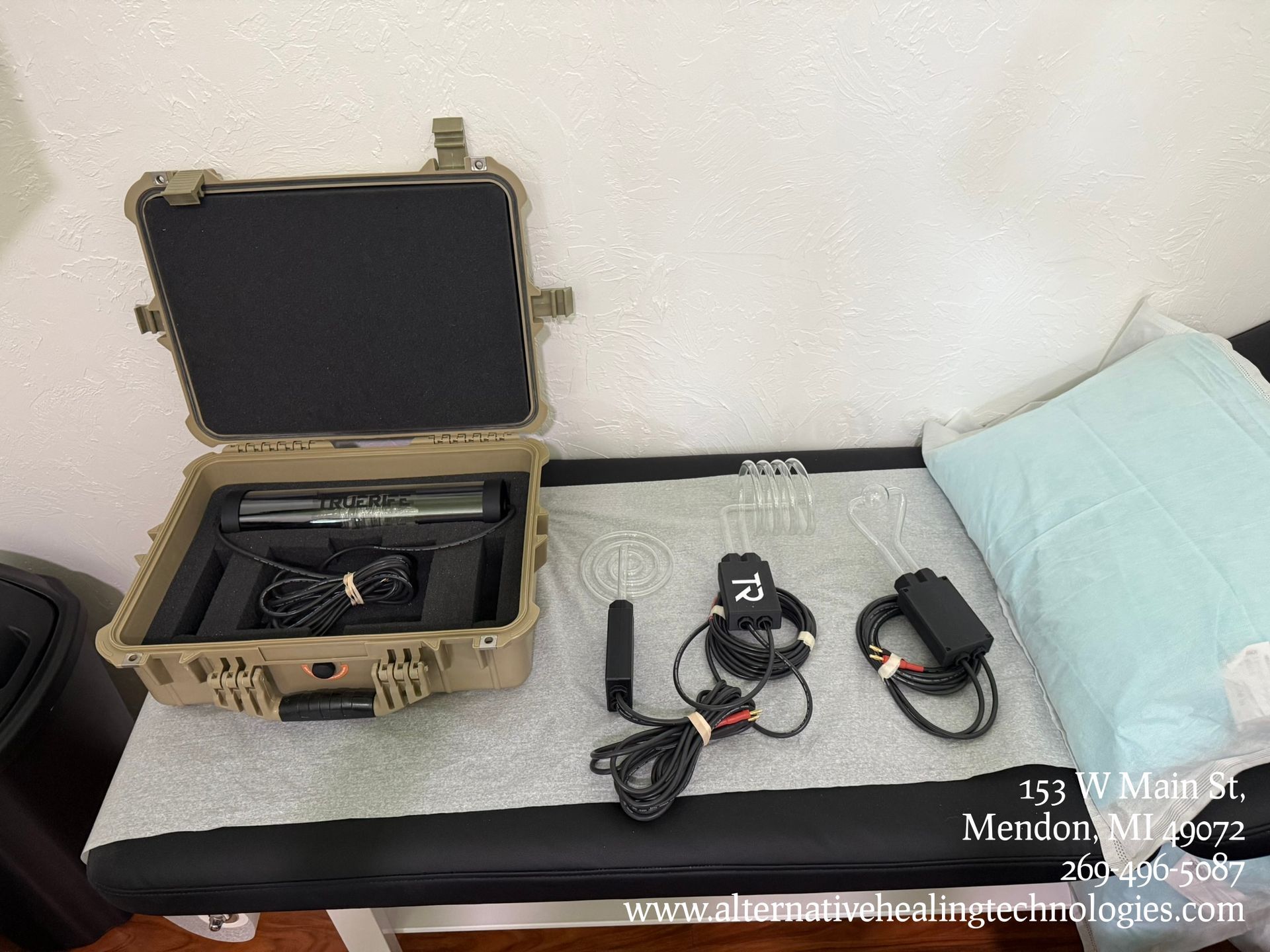 Open tan hard case with camera gear on a bed, beside two black devices and cables on a carpeted floor.