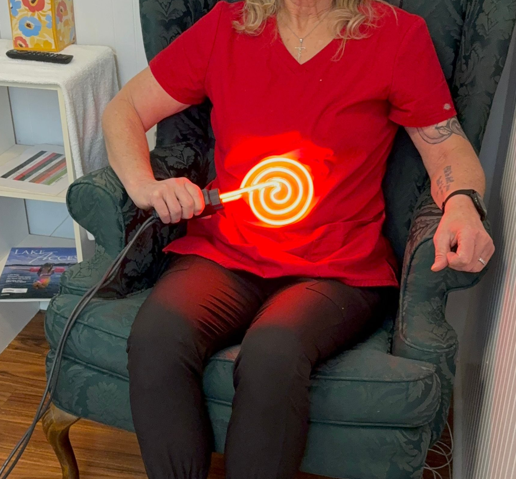 Person seated in a green chair holding a glowing orange spiral true rife light on a red shirt