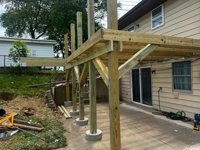 Wooden deck frame under construction beside a house, with posts and concrete footings on a patio.