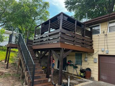 Raised wooden deck with stairs and railing beside a house and garage, surrounded by trees