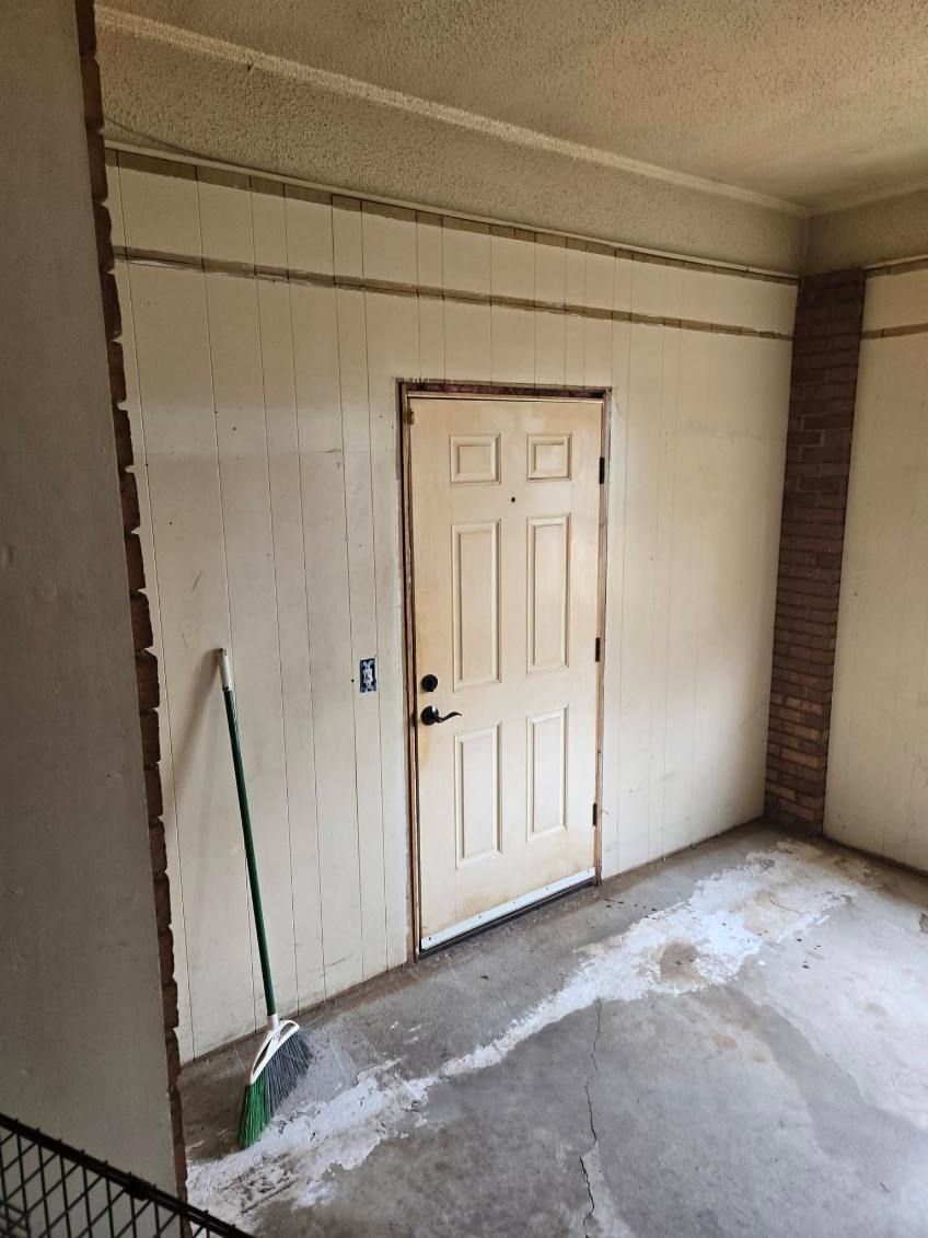 Small unfinished room with a closed beige door, exposed brick corners, and a broom leaning on the left.