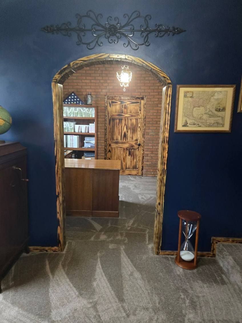 Blue-walled room with arched doorway, wooden shelves, framed map, and an hourglass on a small table.