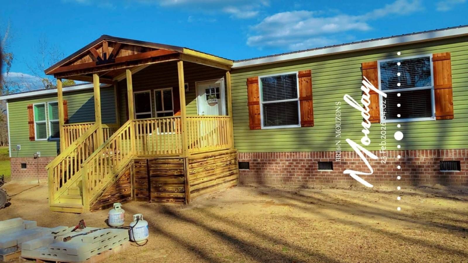 Colorful mobile home with yellow porch on a dirt lot, trees behind, and a small white dog in front.