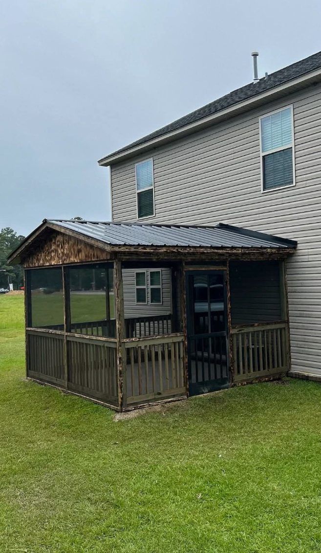 Small weathered wooden porch/shed attached to a gray house on a grassy yard under cloudy sky