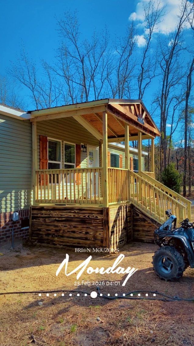 Small house with a wooden porch and stairs in a wooded yard on a sunny day