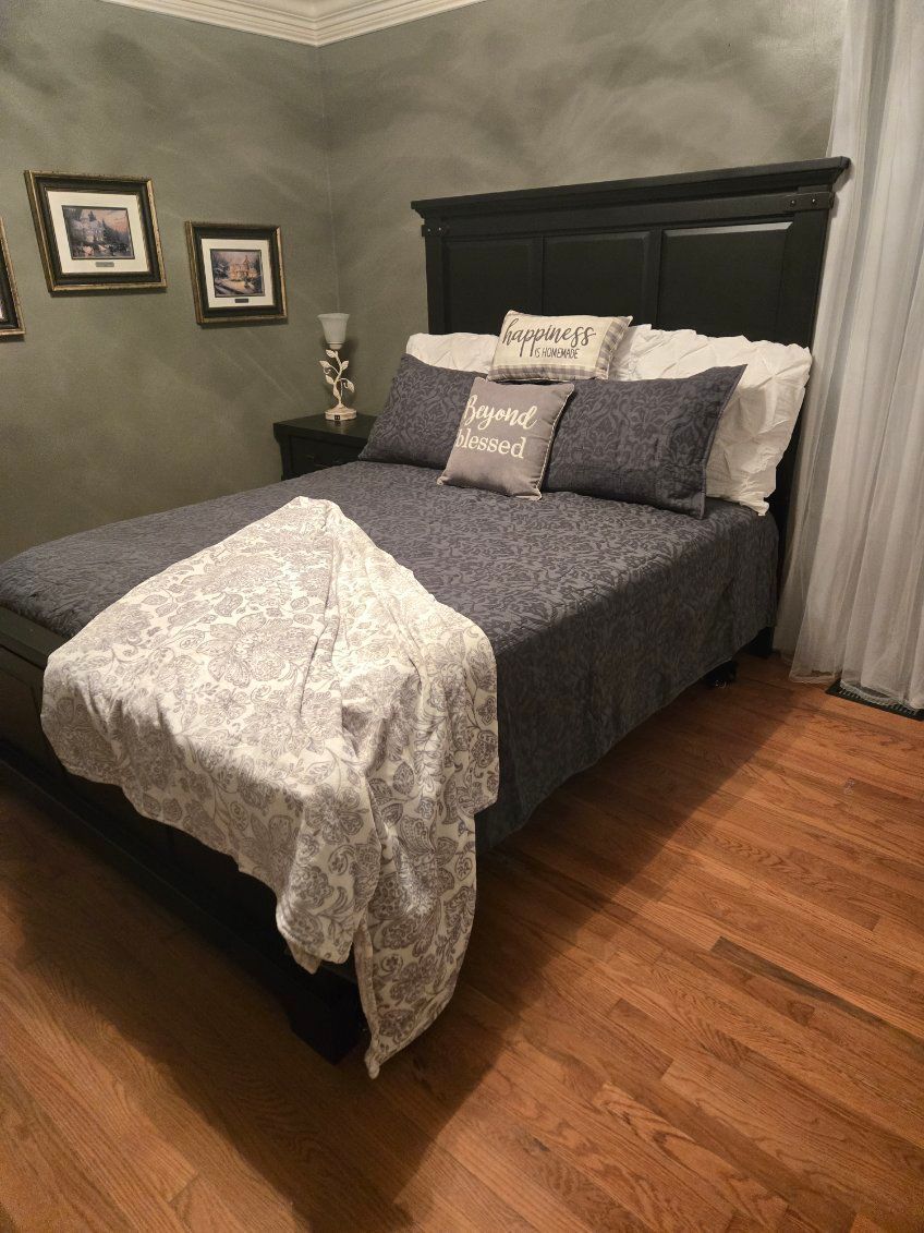 Neatly made bed with dark gray bedding and white throw in a gray-walled bedroom with wood floors.