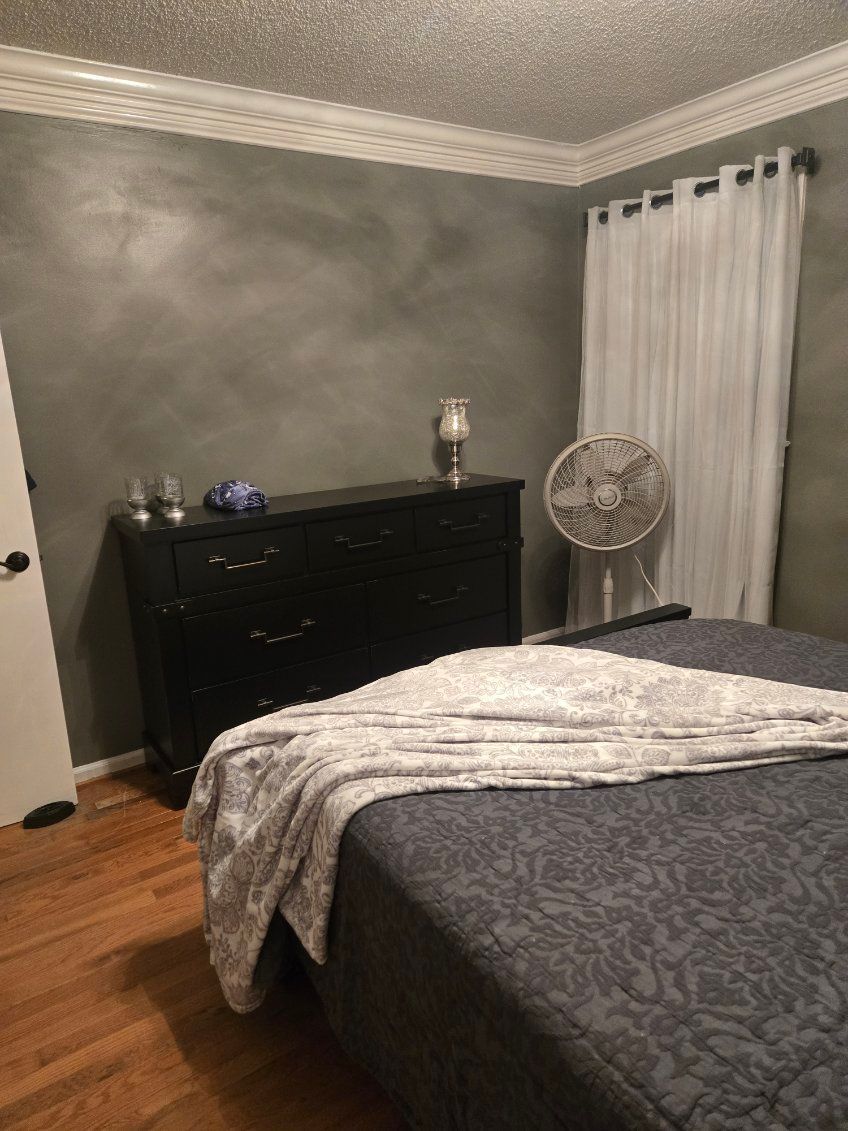 Bedroom with gray bedspread, black dresser, fan, and white curtains against gray walls.
