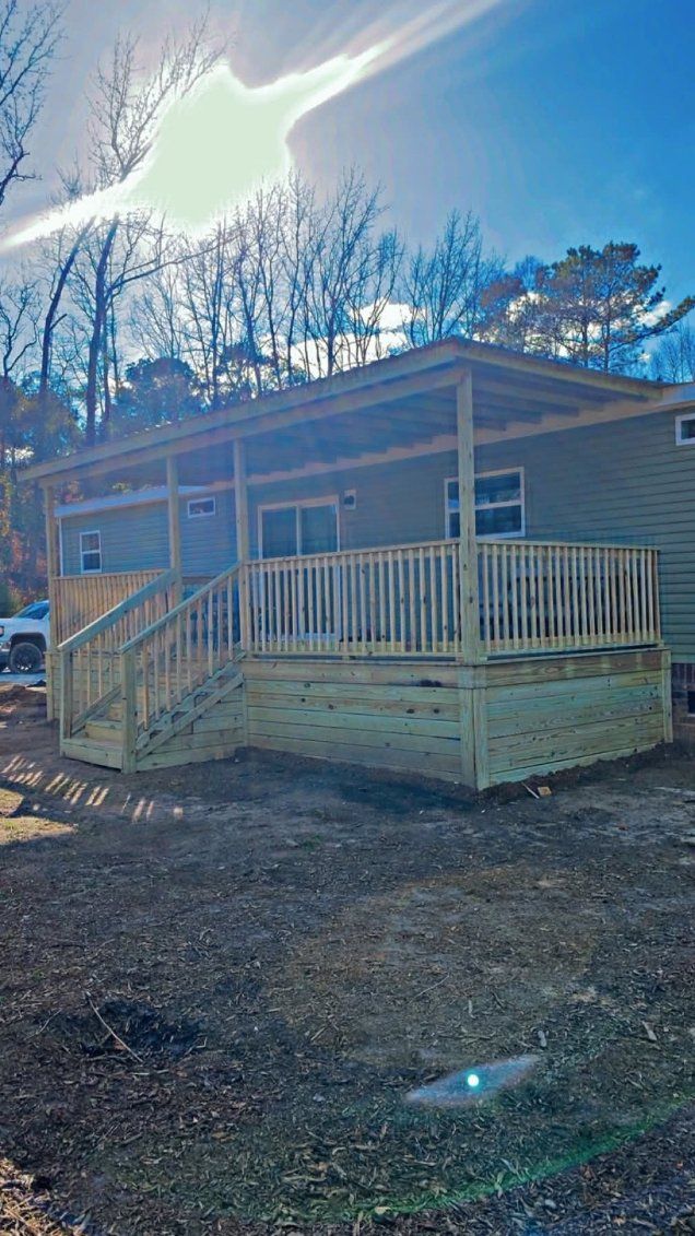 Small tan house with a wooden porch and railing under a bright sun in a wooded yard