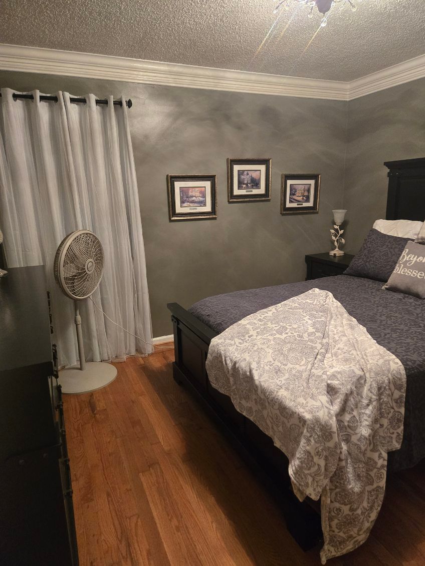 Gray bedroom with a bed, framed wall art, curtains, standing fan, and hardwood floor.