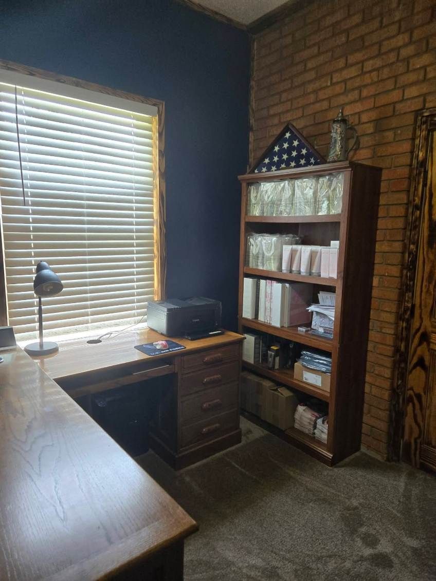 Home office with wood desk, lamp, bookshelf, window blinds, and brick wall.