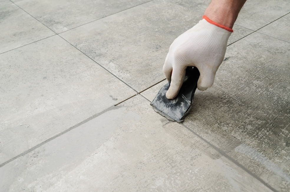 A Hand In A White Work Glove Uses A Tool To Apply Grout Between Light Gray Floor Tiles — HJK Plastering In Kyogle, NSW