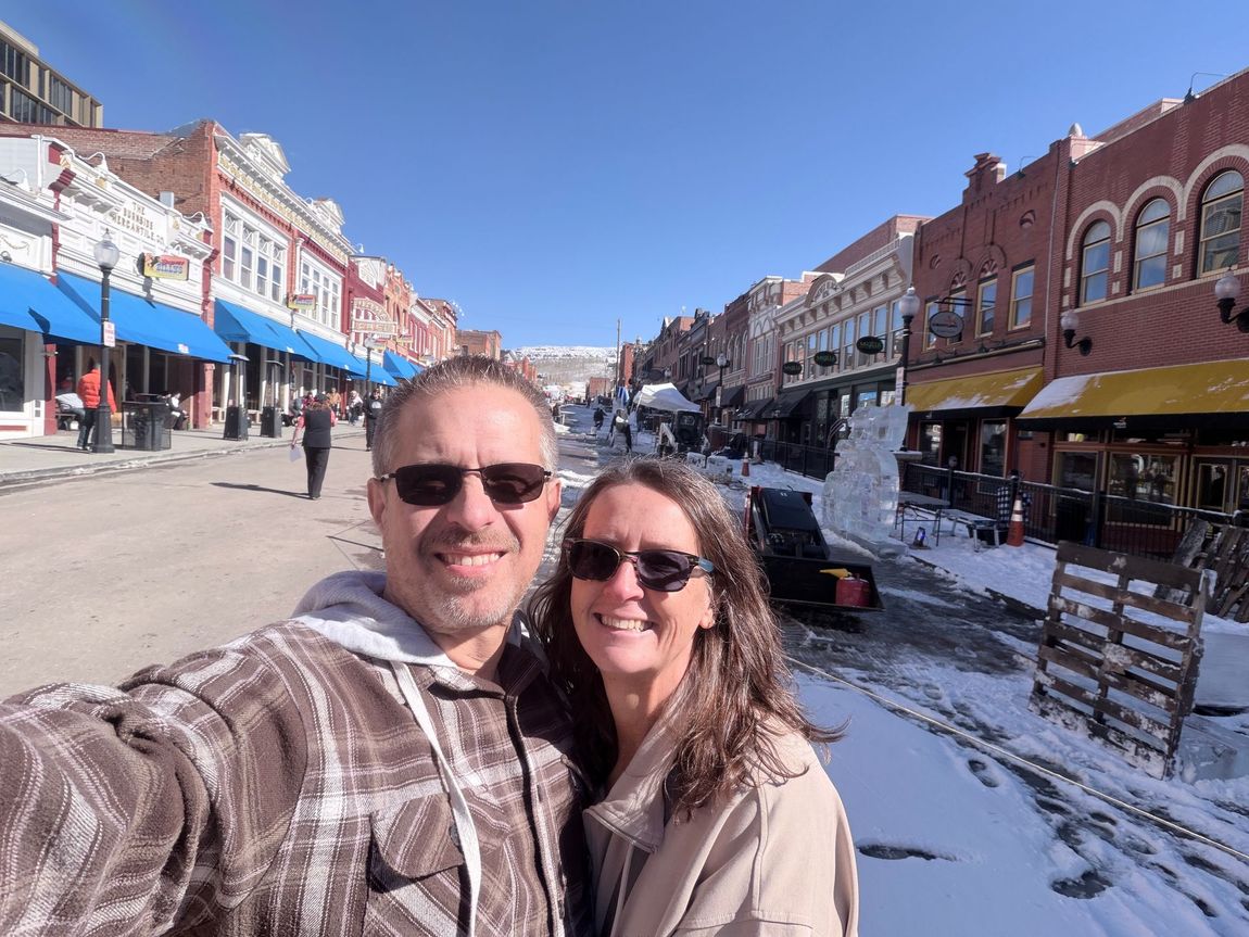 Smiling couple taking a selfie on a snowy main street lined with brick storefronts under a blue sky