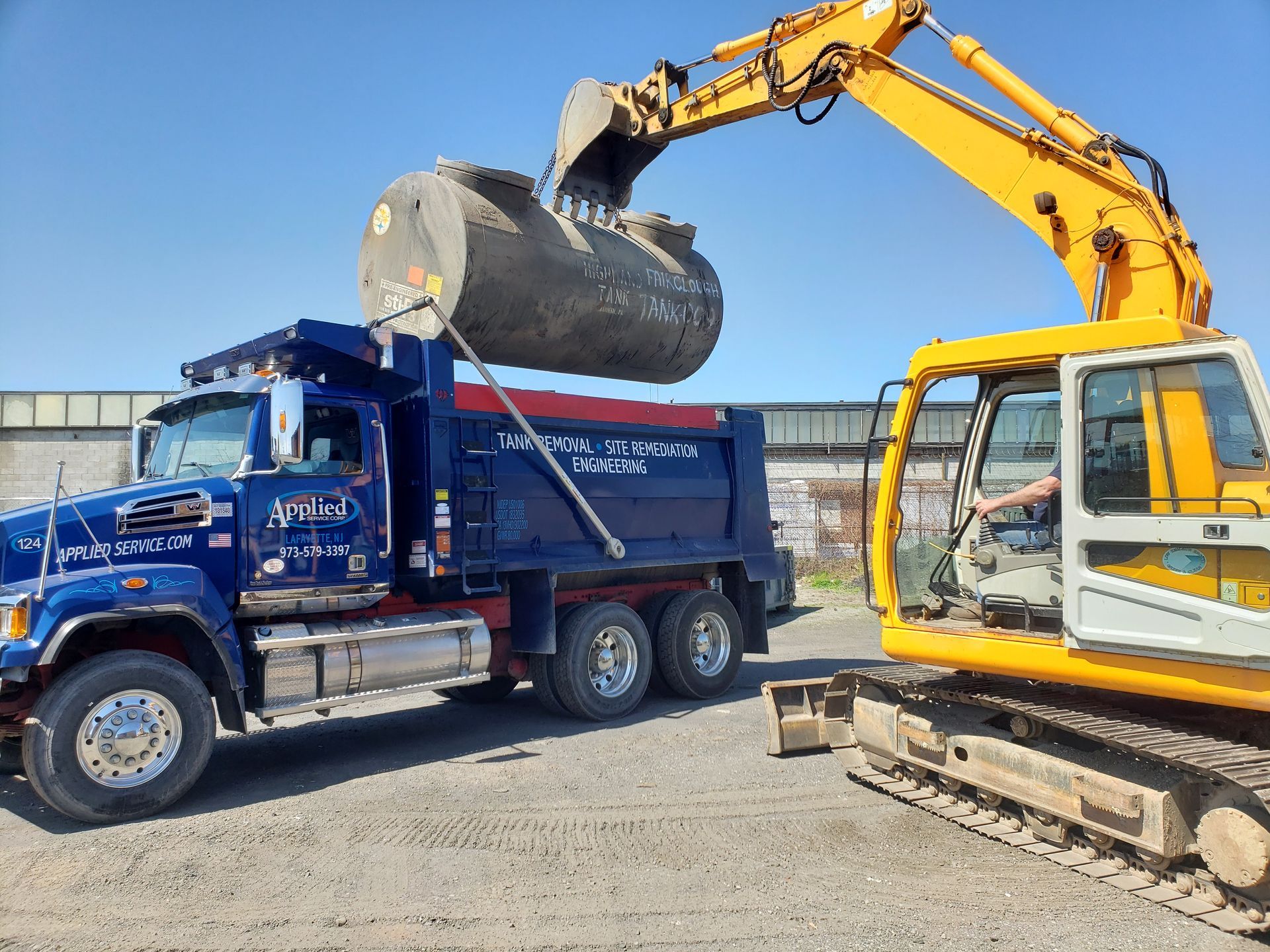 A yellow excavator loading a large, gray object into a blue dump truck on a gravel lot.