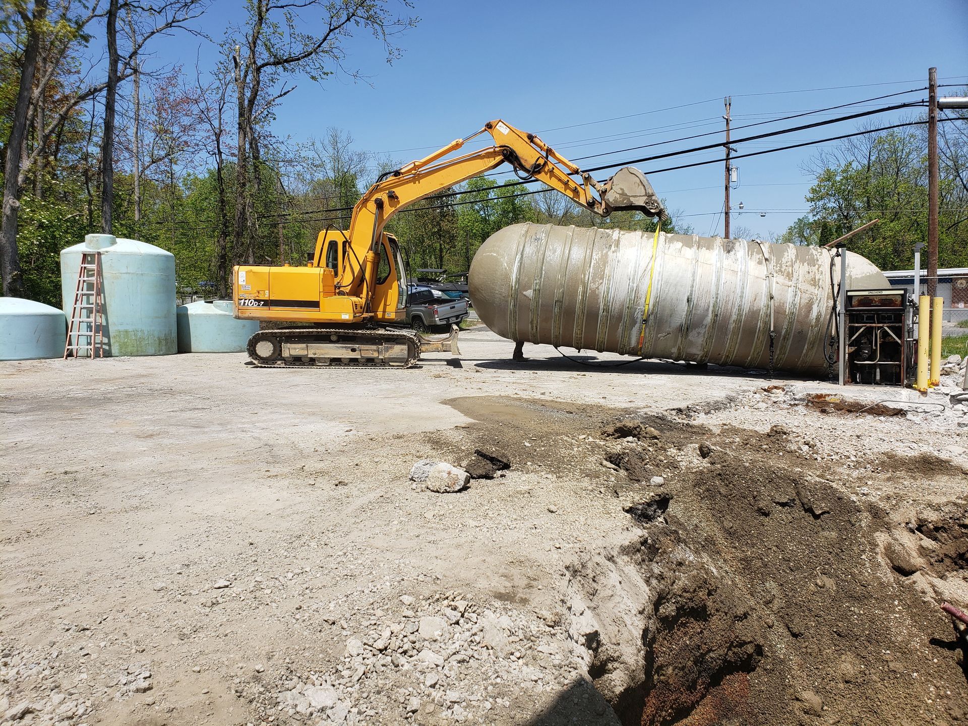 Yellow excavator lifts dirt from a large, white tank on a gravel surface.