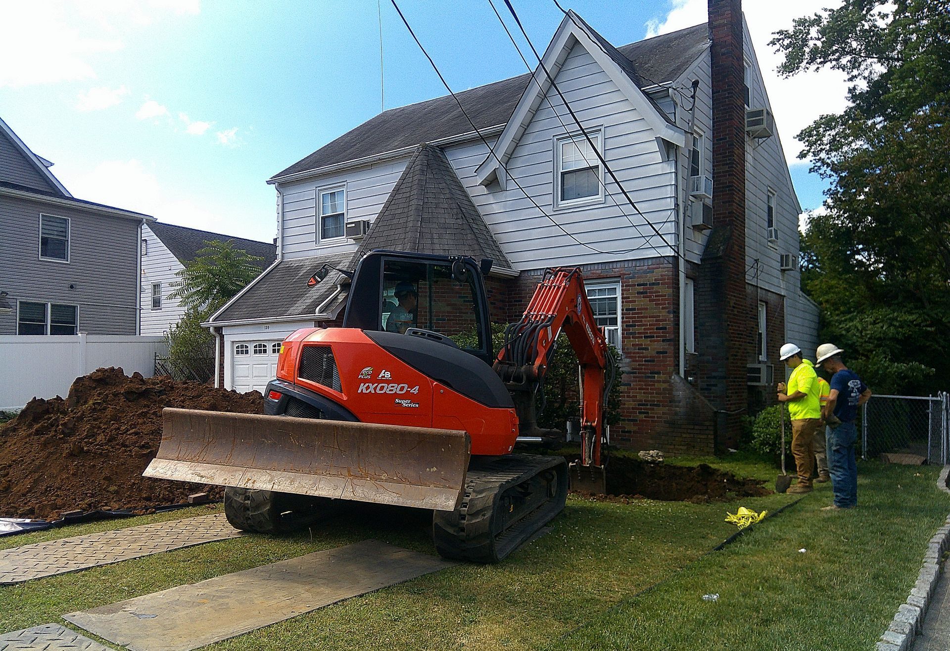 Orange excavator digging near a house, two workers watching on a sunny day.