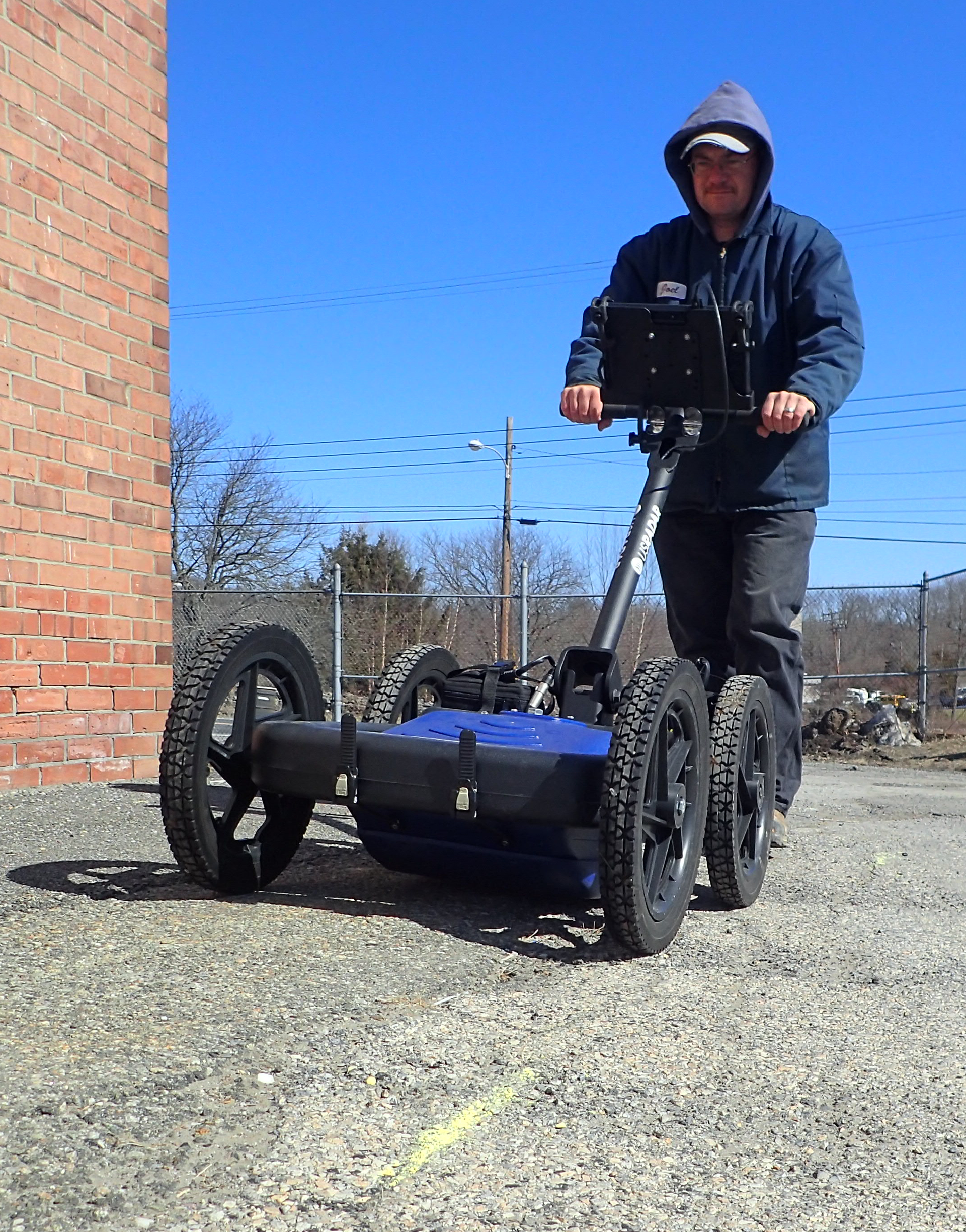 Man operating a ground-penetrating radar on a gravel surface near a brick building under a blue sky.
