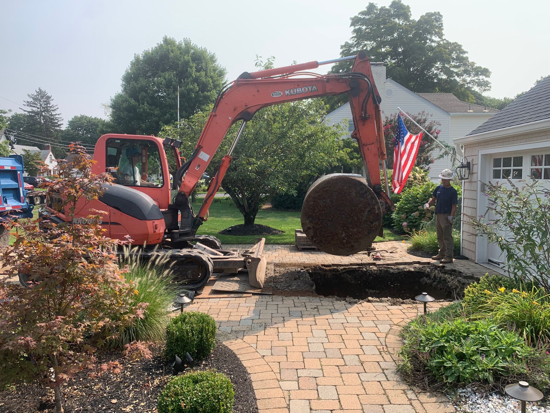 Excavator digging near a brick walkway and house, with an American flag.
